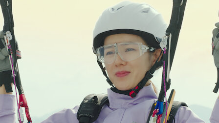Nossa protagonista está suspensa no parapente, usando óculos, capacete e macacão de proteção. O parapente é como um "paraquedas" com umas alças nas quais o condutor pode direcionar seu voo. 