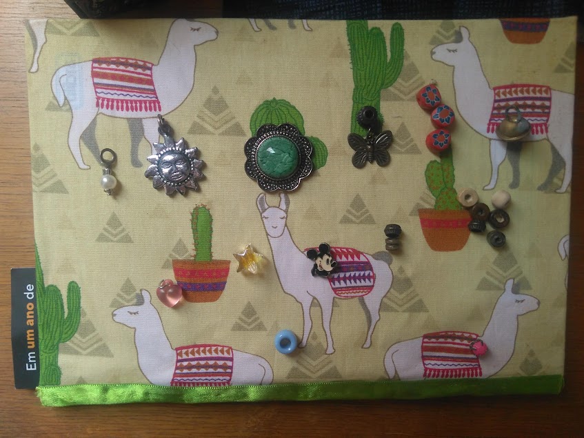 Capa de tecido do meu caderno (estampa de lhamas e cactos). em cima da capa, coloquei algumas coisinhas que achei em meu guarda-roupa e não uso mais. São pingentes diversos: sol, imitação de uma pedra verde, miçangas circulares, mickey, coração e estrelha de plástico, guizo, pérola de imitação, etc.