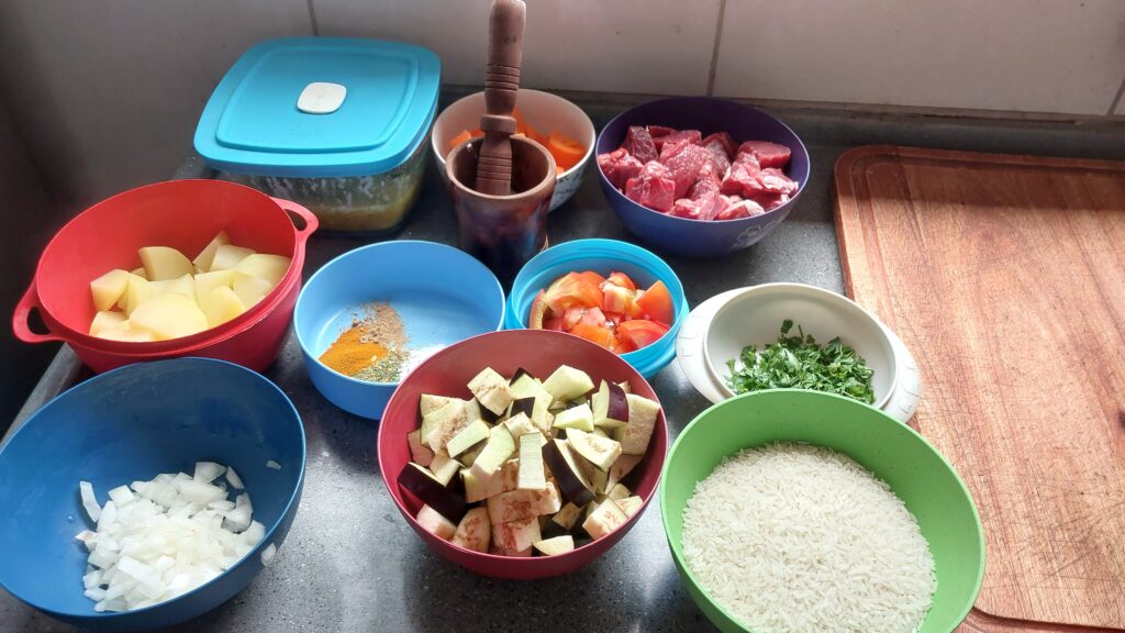 Ingredientes diversos separados em diferentes recipientes. Temos carnet, tomate, batata, cebola, temperos diversos, cenoura e um pilão onde amassei alho. Ao lado direito, uma tábua de madeira,